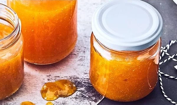 Ein Glas hausgemachte Pfirsichmarmelade neben frischen Pfirsichen.